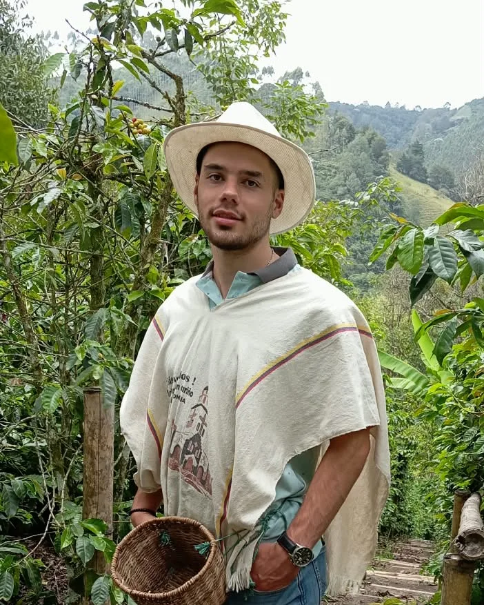 Marco Rico among coffee plants, wearing a hat and a traditional poncho and holding a harvest basket.